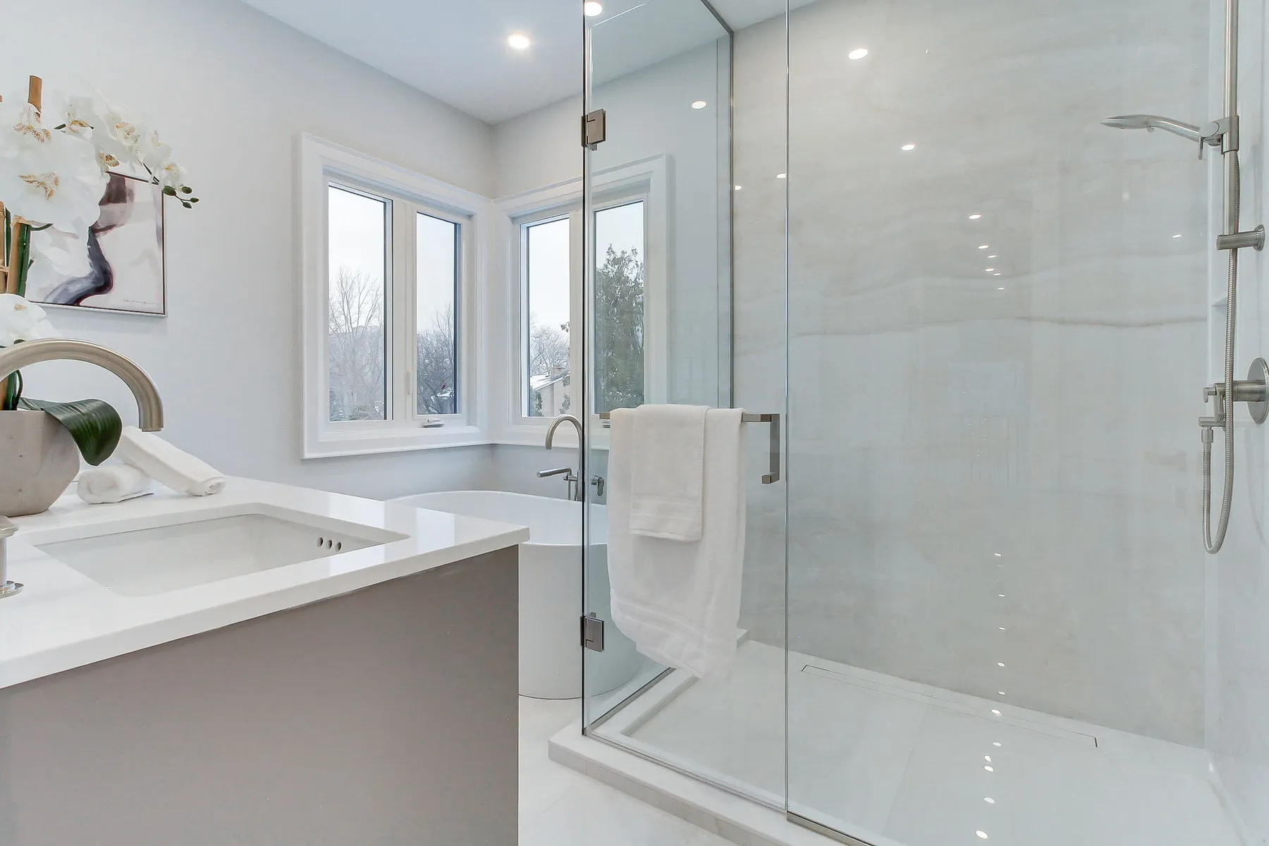 A bright bathroom with a white quartz vanity, a freestanding soaker tub and a glass-enclosed marble shower