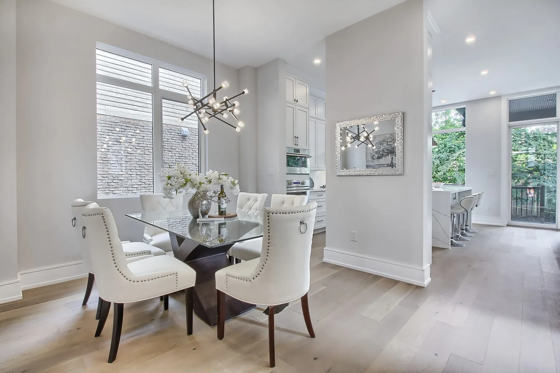 A dining area with a glass-top table, white tufted nailhead chairs and a black sputnik chandelier by large windows