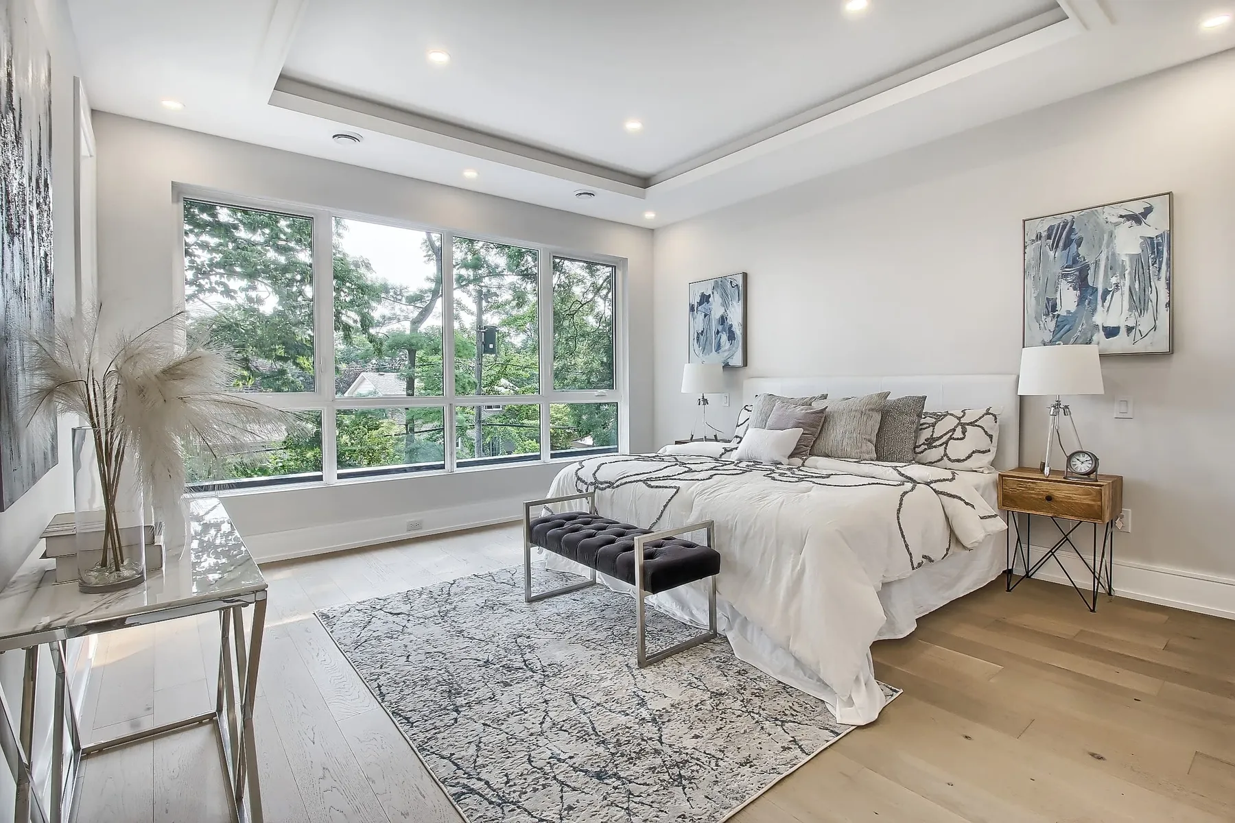 A primary bedroom with a king bed in white linens, a tray ceiling and a wide window framing green foliage