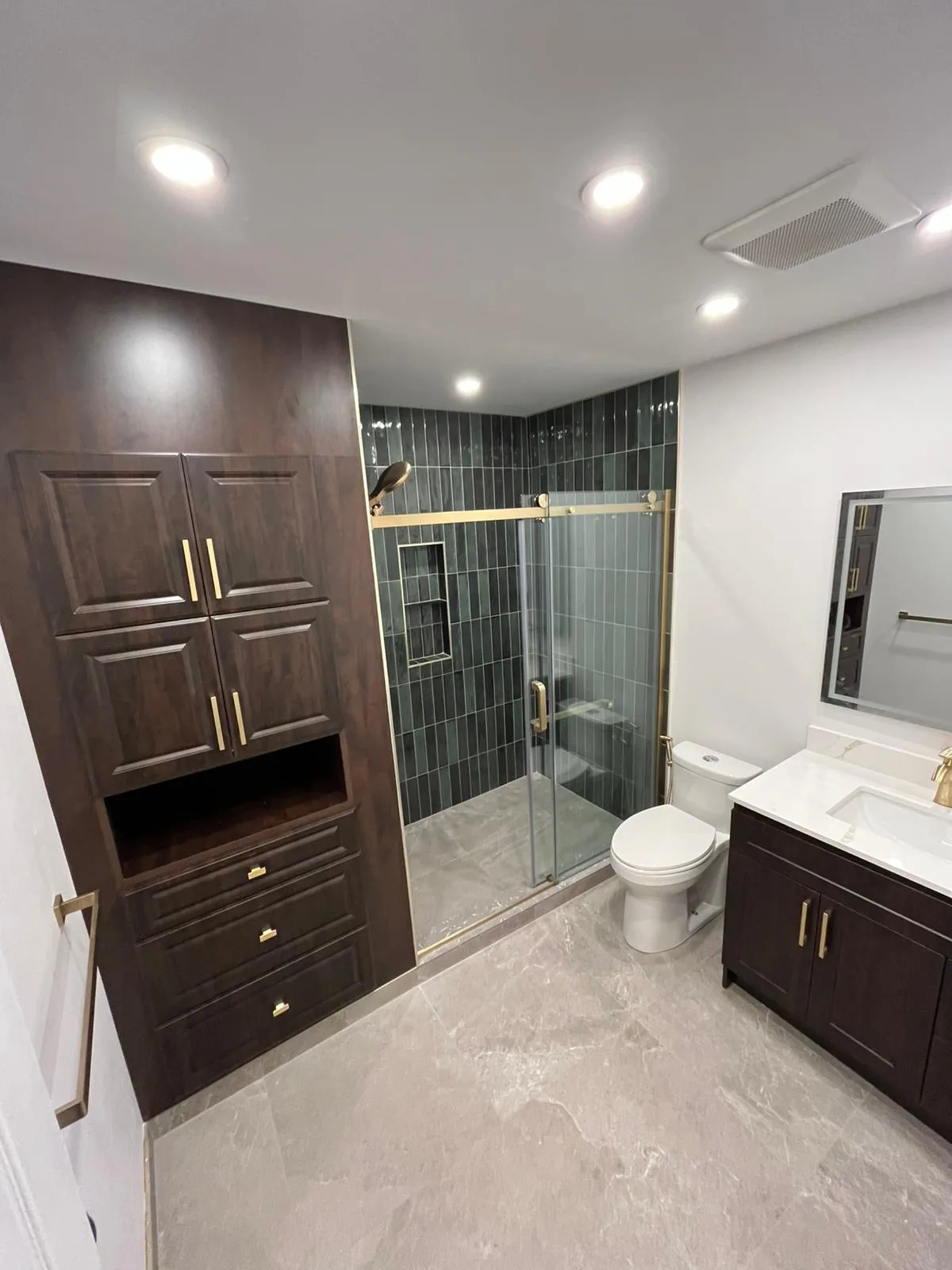 A renovated bathroom with a green-tiled glass walk-in shower, brass fixtures and walnut storage